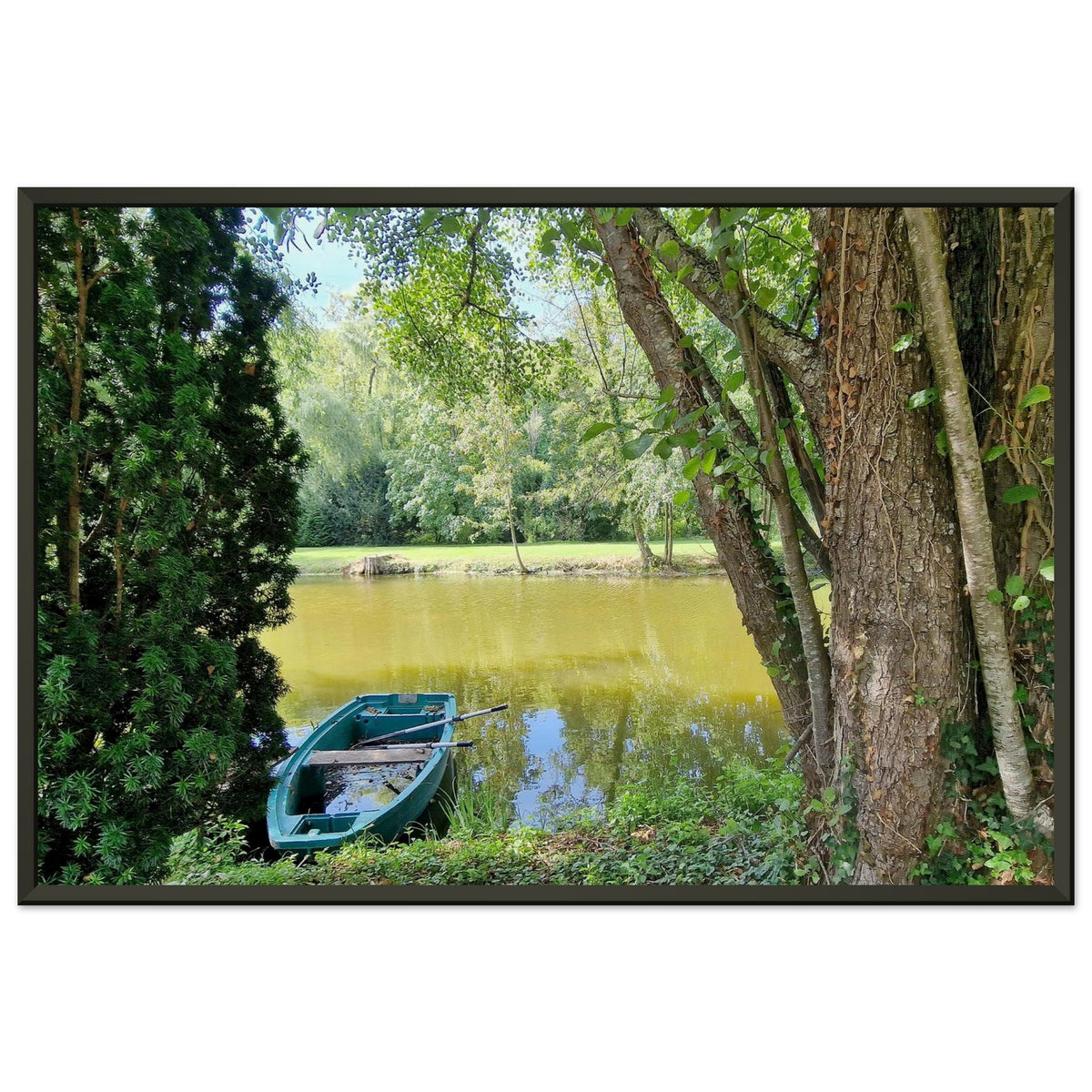 Petite barque en plastique bleu avec ses rames, amarrée au bord d'un étang paisible entouré d'arbres Photo d'art avec cadre