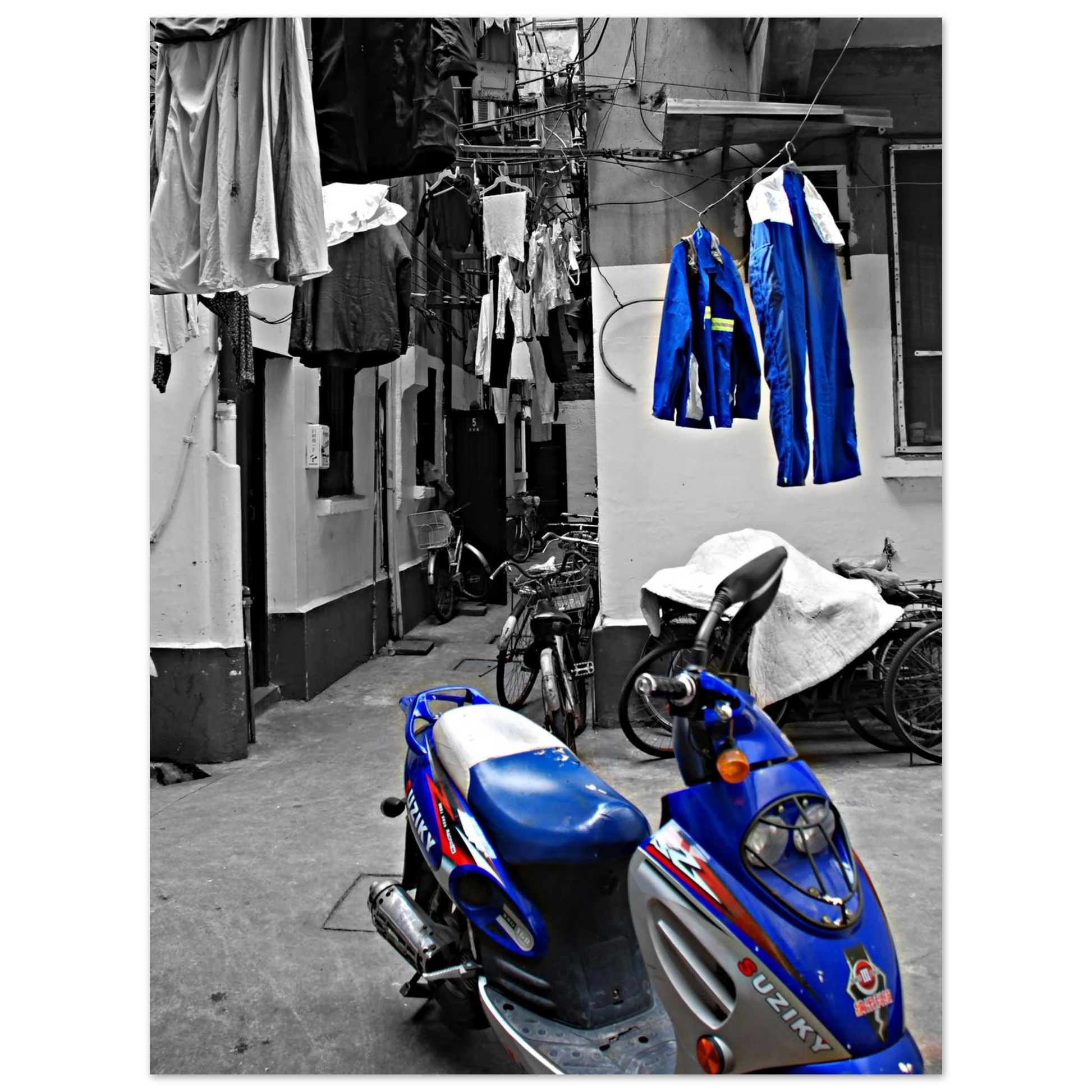 Photographie d'art en noir et blanc d'une ruelle typique de Shanghai avec un scooter bleu vif de marque Suziky et du linge suspendu en couleur sélective.